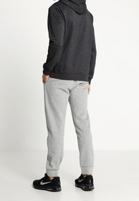 Person som står med händerna i fickorna iförd mörkgrå hoodie, ljusgrå sweatpants och svarta Nike Air Max sneakers.