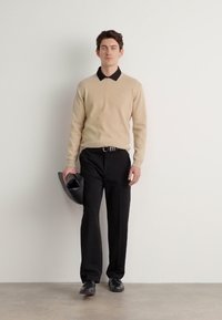 Beige kötött pulóver fekete galléros inggel alatta, fekete nadrággal párosítva. A modell egy fekete kalapot tart a kezében, fekete cipőt visel.