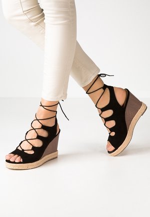 Sorte wedgesandaler i ruskind med udskåret design og krydset snøredetalje. Naturlig tekstureret kilehæl med tovdetaljer. Åben tå-design.