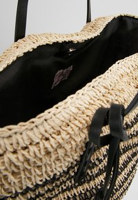 Tkaná slámová kabelka s černými koženými akcenty, s kulatým otvorem, vnitřním podšívkou a malou značkovou visačkou uvnitř.