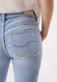 Light blue denim jeans with a mid-rise waist, featuring a back pocket with a subtle distressed detail and a leather brand patch.
