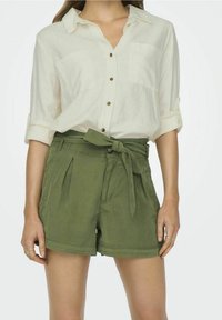 Chemise beige clair à boutons, avec des manches retroussées et deux poches avant, associée à un short ceinturé vert olive avec des plis.