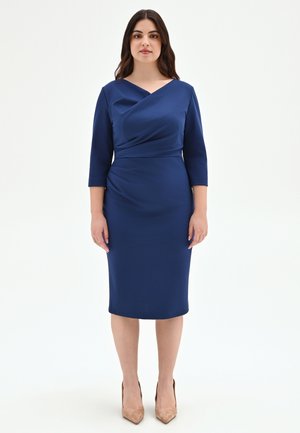 Femme debout, de face, portant une robe bleue marine ajustée arrivant aux genoux avec des manches trois-quarts et des talons hauts beige.