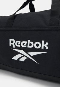Geantă de voiaj neagră cu o textură netedă, având un logo alb proeminent Reebok și închidere cu fermoar. Mâini duble robuste pentru un transport ușor.