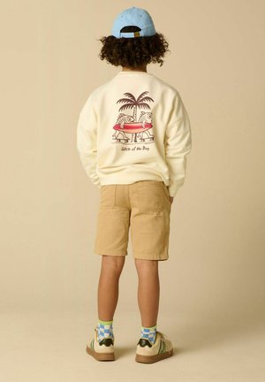 Enfant portant un short beige, un sweat crème avec un imprimé poissons et palmiers, une casquette bleue portée à l'envers, des chaussettes à motifs et des sneakers blanches, debout de dos.