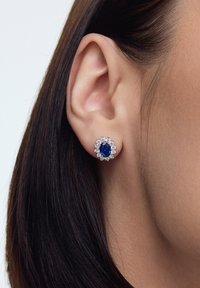 Niebieskie kolczyki z owalnym kamieniem szlachetnym, z okrągłymi przezroczystymi akcentami, osadzone w srebrze, posiadają misterny wzór kwiatowy wokół centralnego kamienia.