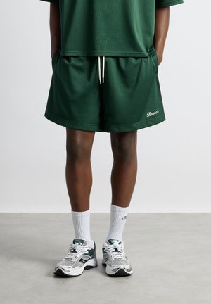 Une personne porte un short en mesh vert foncé avec un cordon blanc, un haut assorti, des chaussettes blanches et des chaussures de course blanches et vertes, debout sur un sol gris.
