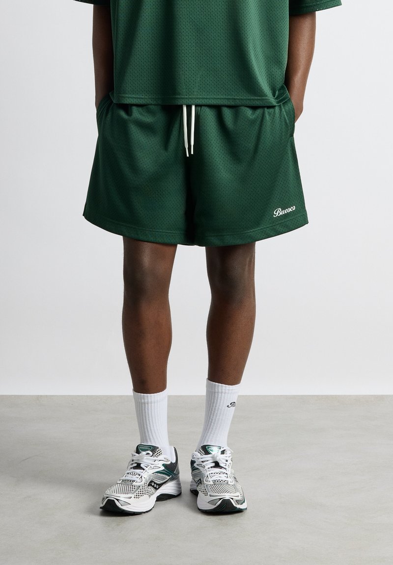 Une personne porte un short en mesh vert foncé avec un cordon blanc, un haut assorti, des chaussettes blanches et des chaussures de course blanches et vertes, debout sur un sol gris.
