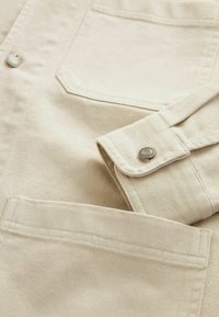 Camicia di cotone beige con chiusura a bottoni, dotata di tasca e tessuto a trama. Il colletto e i polsini sono piegati con cura. Strisce orizzontali sottili.