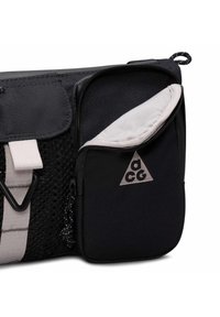 Sac en tissu noir et gris avec une poche frontale partiellement ouverte affichant un intérieur gris clair et un logo triangulaire "acg".