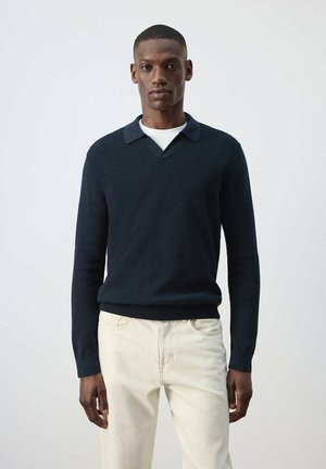 Hombre con un suéter de manga larga azul oscuro con cuello sobre una camisa blanca y pantalones beige claros, de pie frente a un fondo claro y liso.