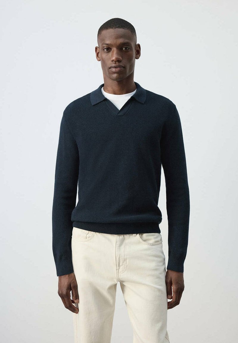 Homme portant un pull bleu foncé à manches longues avec col par-dessus une chemise blanche et un pantalon beige clair, debout devant un fond clair uni.