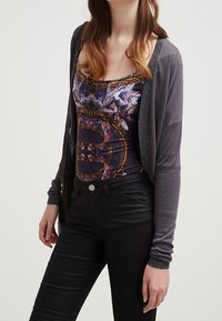 Grå cardigan med glittrande detaljer, bärs över en tanktop med flerfärgad mönster. Svarta åtsittande jeans fulländar outfiten.