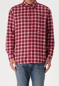 Mężczyzna ubrany w czerwoną, czarną i białą koszulę w kratę z długimi rękawami oraz niebieskie dżinsy, stojący na jednolitym tle.