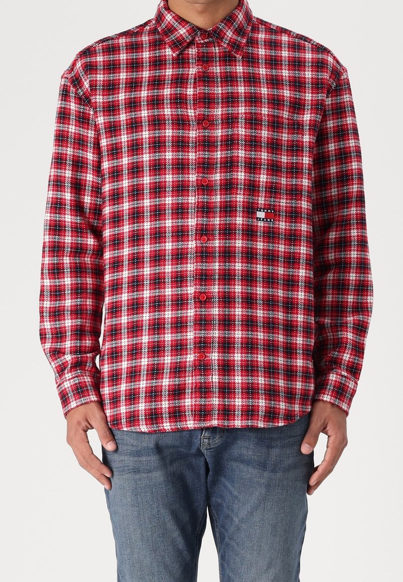 Mężczyzna ubrany w czerwoną, czarną i białą koszulę w kratę z długimi rękawami oraz niebieskie dżinsy, stojący na jednolitym tle.