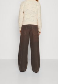 Weite braune Hose mit leicht strukturierter Oberfläche, ausgestattet mit Gesäßtaschen und kontrastierenden Nähten. Kombiniert mit einem leichten, gemusterten Pullover.