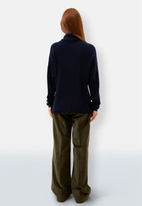 Marine-blaues Strickpullover mit hohem Kragen, weicher Textur und entspannter Passform, kombiniert mit olivgrünen, locker sitzenden Hosen aus leichtem Stoff.