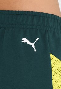 Primer plano de pantalones cortos deportivos de color verde oscuro con cinturilla elástica, logo blanco de Puma y panel lateral de malla amarilla.