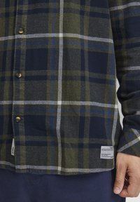 Camisa de franela a cuadros en verde, azul marino y blanco con botones marrones, con un dobladillo redondeado y una etiqueta tejida en la esquina inferior derecha.