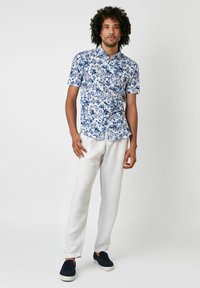 Kurzärmliges Blumenhemd in Weiß und Blau mit Knopfleiste; kombiniert mit hellen Leinenhosen und schwarzen Slippern.