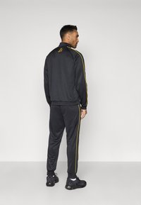 Čierny športový trenčkot s žltými bočnými pruhmi. Bunda má vysoký golier a priliehavé manžety. Nohavice majú elastické členky.