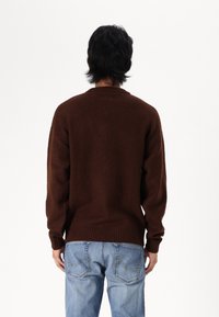 Hnědý pletený svetr s kulatým výstřihem, žebrovanými manžetami a lemem, nošený přes světle modré džíny. Tkanina má měkký, texturovaný vzhled.