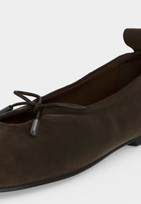 Σκούρο καφέ σουέτ επίπεδο παπούτσι με στρογγυλεμένη μύτη, λεπτομέρεια φιόγκου με μεταλλικά άκρα και χαμηλή μαύρη σόλα, εμφανιζόμενο από κοντινή γωνία.