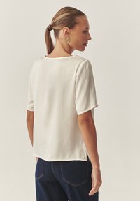 Blusa de manga corta color crema, hecha de una tela suave, con escote redondeado y aberturas laterales. Combinada con jeans oscuros.