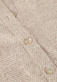 Beige textielstof met een visgraatpatroon, voorzien van pailletten en twee lichte, ronde knopen. Glad afwerking.