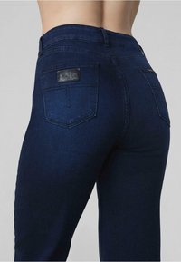 Jeans de denim azul oscuro con un diseño ajustado, estilo de cinco bolsillos y un parche de logo de cuero en el bolsillo trasero.