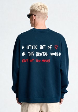 Persona con capelli biondi corti e occhiali da sole indossa una felpa blu navy con il testo "A little bit of heart in this brutal world (but not too much)" sulla schiena.
