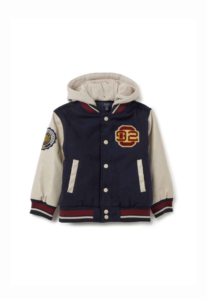 Chaqueta varsity azul marino con capucha crema, detalles a rayas rojas y blancas, logo bordado y dos bolsillos frontales. Textura de tela suave.