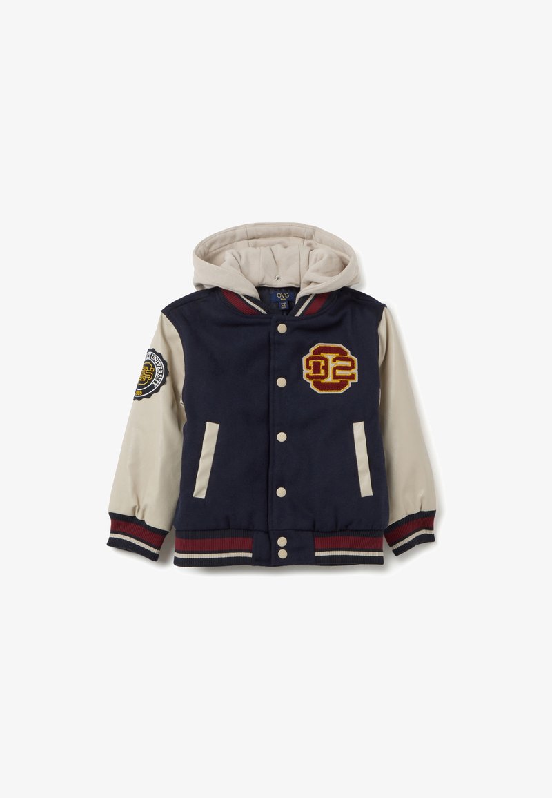 Chaqueta varsity azul marino con capucha crema, detalles a rayas rojas y blancas, logo bordado y dos bolsillos frontales. Textura de tela suave.