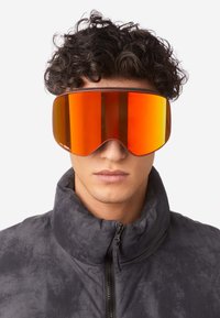 Occhiali da sci con una grande lente arancione specchiata, montatura nera e un design elegante; indossati da una persona in una giacca bomber scura e strutturata.