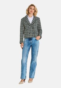 Frau, die ein schwarz-weiß kariertes, doppelt geknöpftes Blazer, eine weiße Bluse, helle Blue Jeans und beige High Heels trägt, steht mit einer Hand in der Tasche.