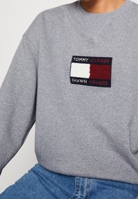 Šedá mikina s kulatým výstřihem, zdobená vyšívanou nášivkou s logem v červené, bílé a tmavě modré barvě. Materiál má jemnou strukturu.