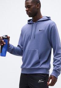 Homme en sweat à capuche Nike bleu et short Nike Pro noir tenant une bouteille d'eau sportive bleue sur fond blanc.