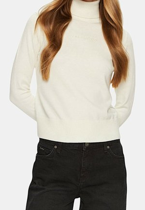 Kobieta w białym swetrze z golfem z napisem „Guess Jeans” na piersi oraz czarnych, wysokich dżinsach, z rękami za plecami.