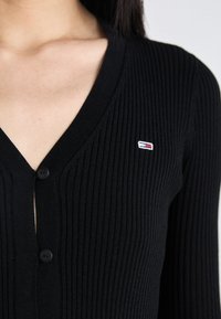 Melna ribota kardigāna ar V veida kaklu, ar pogu aizdari un mazu izšūtu logotipu sarkanā, baltā un zilā krāsā krūtīs.