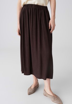 Donna che indossa una gonna plissettata di colore marrone scuro, lunga fino alla caviglia, con una fascia elastica in vita e ballerine beige con punta, su uno sfondo semplice.