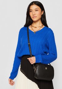 Maglione blu a maglia con scollo a V, abbinato a una gonna a pieghe nera e a una borsa a tracolla nera con accessori in metallo dorato.