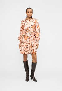 Ženska s pletenimi lasmi, oblečena v svetlo rožnato obleko z dolgimi rokavi in pasnim trakom, obuje temno rjave škornje z visoko peto do kolen.