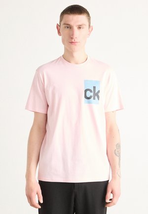 Pink cotton t-shirt featuring a blue rectangular logo with “ck” in bold black letters on the left chest. Short sleeves and crew neckline.