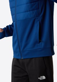 Modrá sportovní bunda s polstrovanou horní částí, texturovanými dolními panely, zipovými kapsami a černými kalhotami s bílým logem na nohavici.