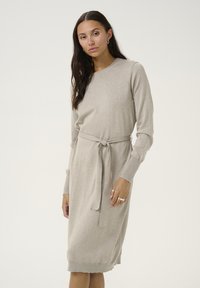 Robe en tricot beige avec des manches longues, un col rond et une ceinture à nouer à la taille. Elle présente une texture lisse et un ourlet légèrement effilé.
