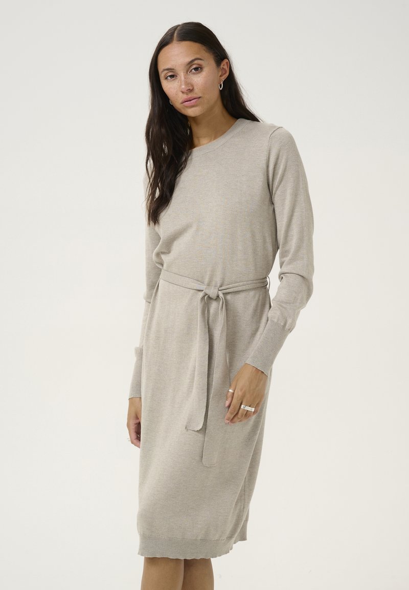 Robe en tricot beige avec des manches longues, un col rond et une ceinture à nouer à la taille. Elle présente une texture lisse et un ourlet légèrement effilé.