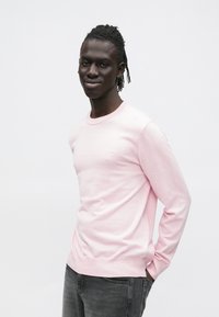 Ροζ ανοιχτόχρωμο πουλόβερ με μακριά μανίκια, στρογγυλό γιακά, ριμπ μανσέτες και τελείωμα. Μικρό λογότυπο στο στήθος, συνδυασμένο με σκούρο γκρι τζιν. Ομαλή υφή.