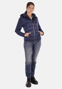 Giacca puffer blu navy con cappuccio, design trapuntato, zip frontale e tasche laterali, abbinata a jeans blu e stivali neri con lacci.