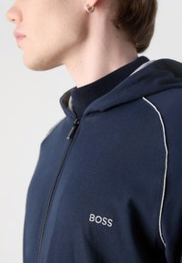 Felpa con cappuccio blu navy in cotone, presenta una chiusura con zip, dettagli in piping bianco e logo ricamato sul lato sinistro del petto.