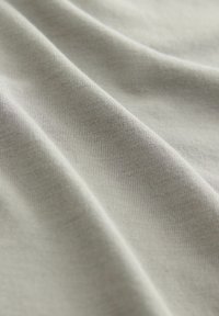 Vaalea harmaa, pehmeä puuvillakangas, jossa on sileä, tekstuurinen pinta, esittäen hellävaraisia taitoksia ja pieniä värin ja valon vaihteluja.